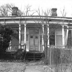 Weathered single-story house with vines growing on covered porch and fence