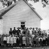 Group of white children and teachers standing outside single-story school house