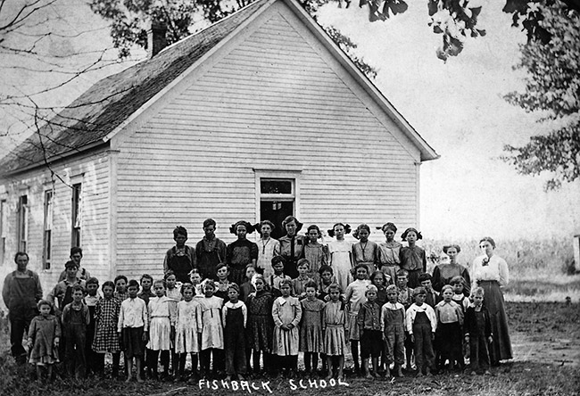 Group of white children and teachers standing outside single-story school house