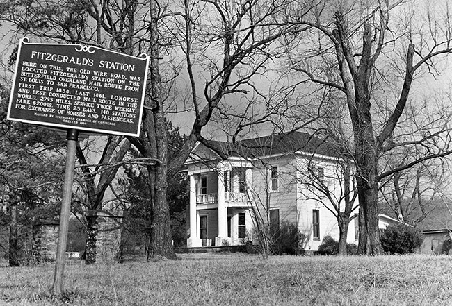 Multistory house with covered porch and balcony under trees with historical marker sign in the foreground