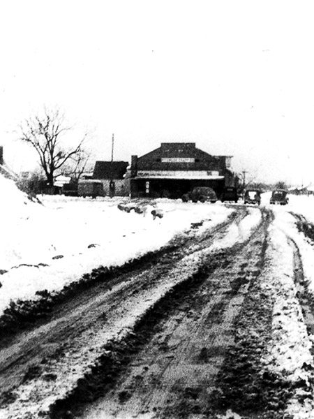 Muddy rutted road with snow alongside leading to brick building with parked cars and bare tree