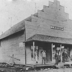 White patrons standing on covered porch of single-story storefront on dirt road