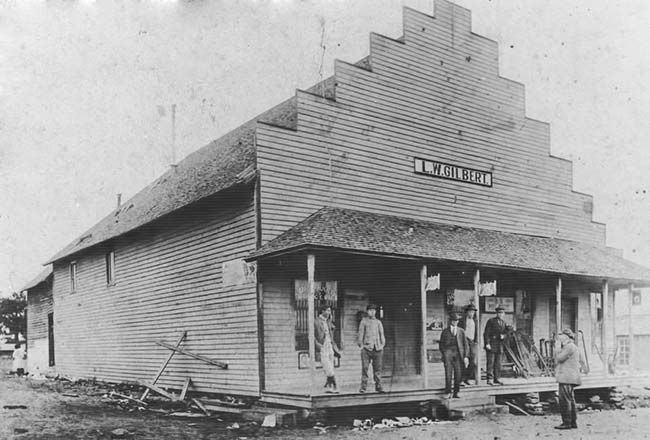 White patrons standing on covered porch of single-story storefront on dirt road