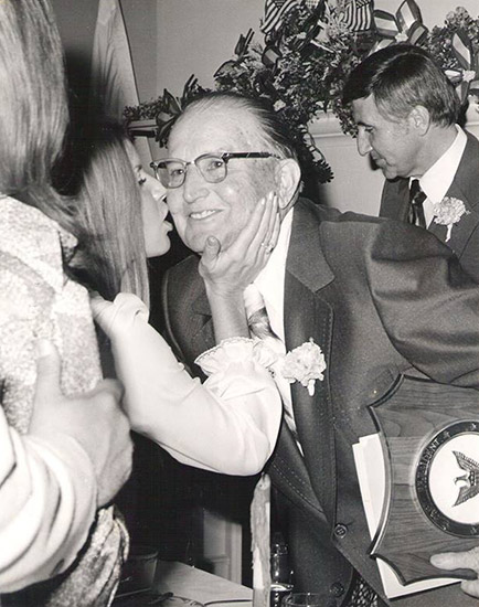 White woman kissing older white man with glasses in suit and tie holding award plaque with white man in suit standing behind him