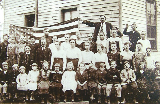 Group of white children and white man holding up an American flag outside school house