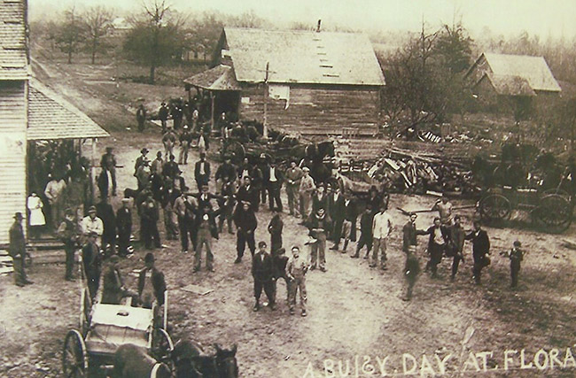 Crowd of people and horse drawn wagons on dirt road with single-story storefronts on both sides