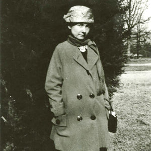 White woman in suit coat and hat standing with tree behind her
