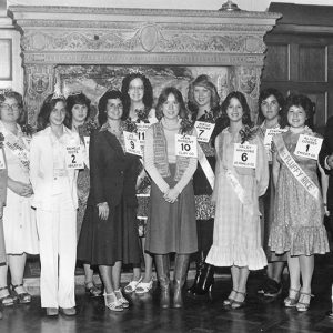 White man in suit and striped tie standing at fireplace with group of white women in dresses with numbered contestant cards