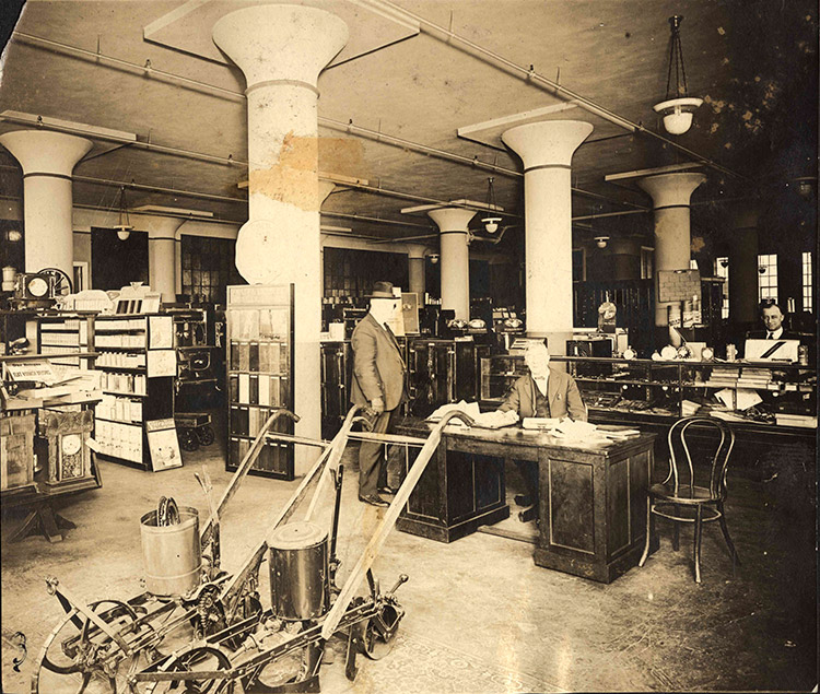 White man in suit and hat standing next to white man sitting at desk with white man standing behind shelf in hardware store