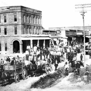 Crowd of people with horse-drawn wagons transporting cotton down a street lined with buildings