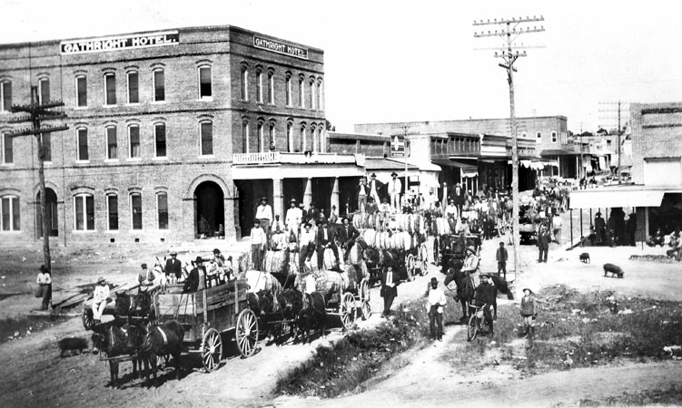 Crowd of people with horse-drawn wagons transporting cotton down a street lined with buildings
