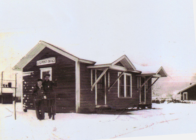 Single-story building with awning and two white people standing in front in snow