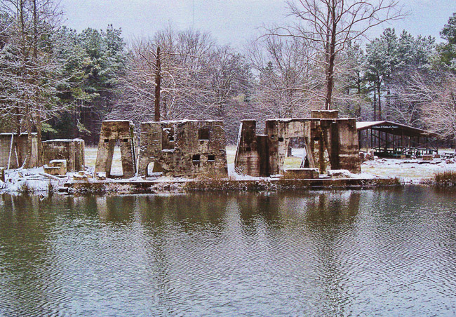 Concrete ruins near edge of pond with pavilion and trees in background during winter