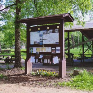 Wooden pavilion in the trees with picnic tables and a bulletin board with signs and maps