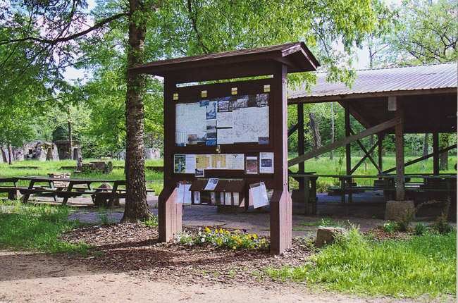 Wooden pavilion in the trees with picnic tables and a bulletin board with signs and maps