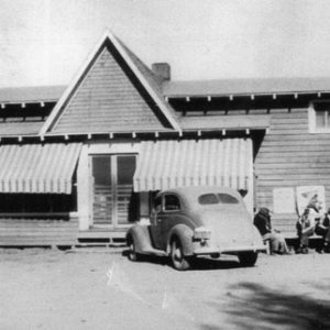 Long building with wood siding and parked car and people sitting in front