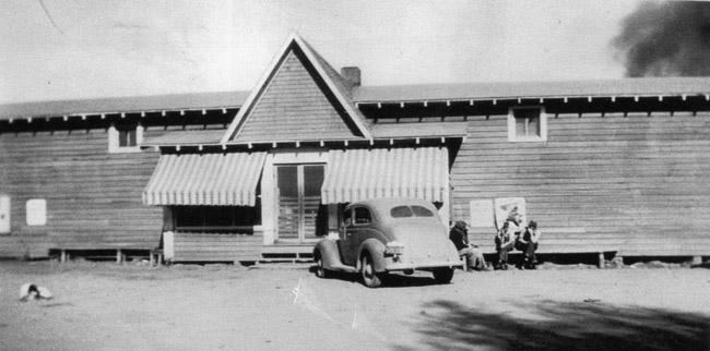 Long building with wood siding and parked car and people sitting in front