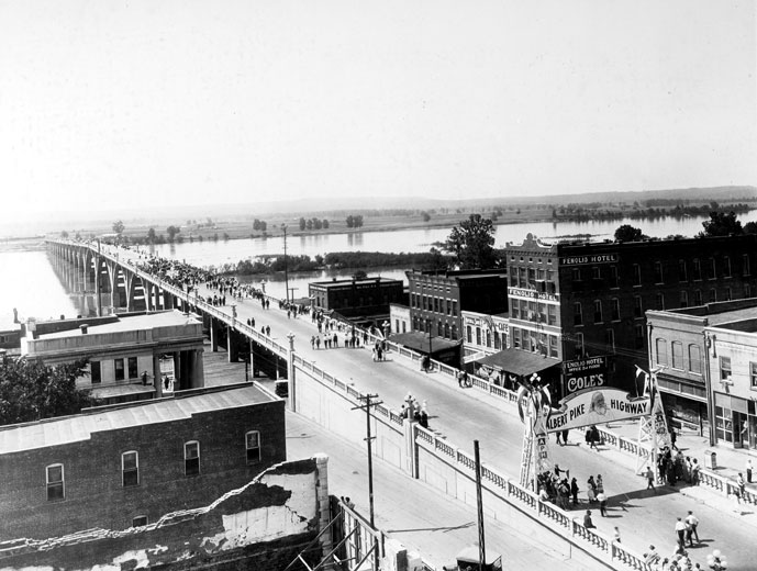 Crowd of people crossing a concrete bridge with buildings in the foreground with sign "Albert Pike Highway"