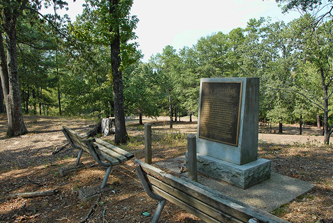 Stone monument with plaque and benches on sloped hill top in forested area