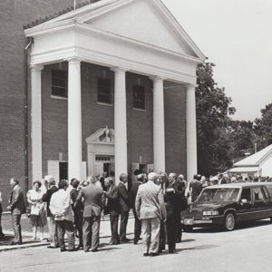 Crowd and hearse outside multistory church building with four columns next to house