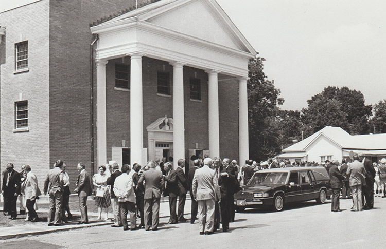 Crowd and hearse outside multistory church building with four columns next to house