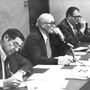 Older white men with glasses in suits sitting at table with microphones