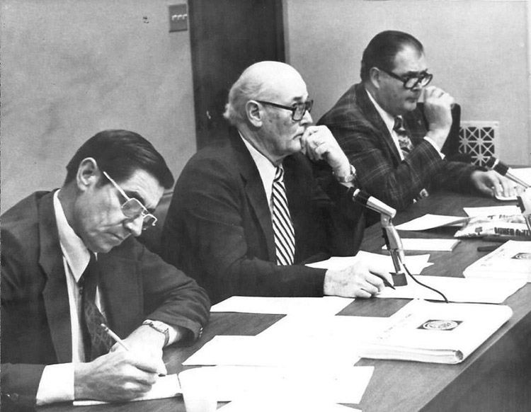 Older white men with glasses in suits sitting at table with microphones