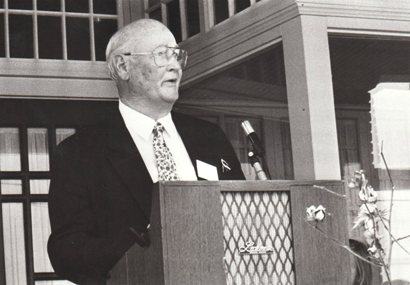 White man in suit and tie speaking into microphone at lectern