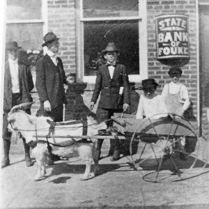 White men and boys standing outside brick building with a goat cart