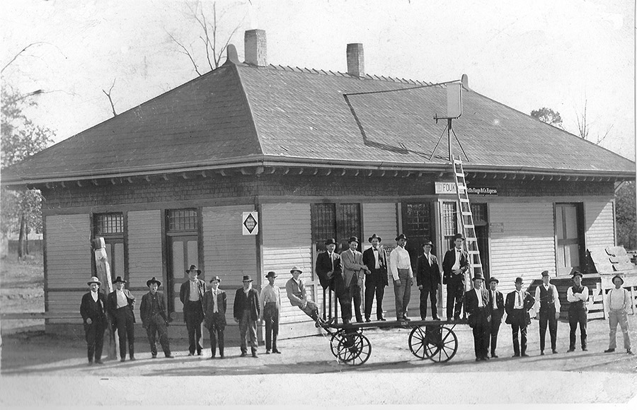 Group of white men and wheeled cart outside railroad depot building