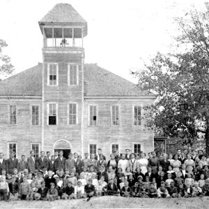 Group of white students and teachers outside multistory school house with bell tower