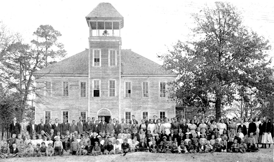 Group of white students and teachers outside multistory school house with bell tower