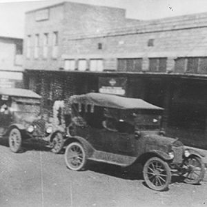 Cars parked outside brick storefronts on dirt road