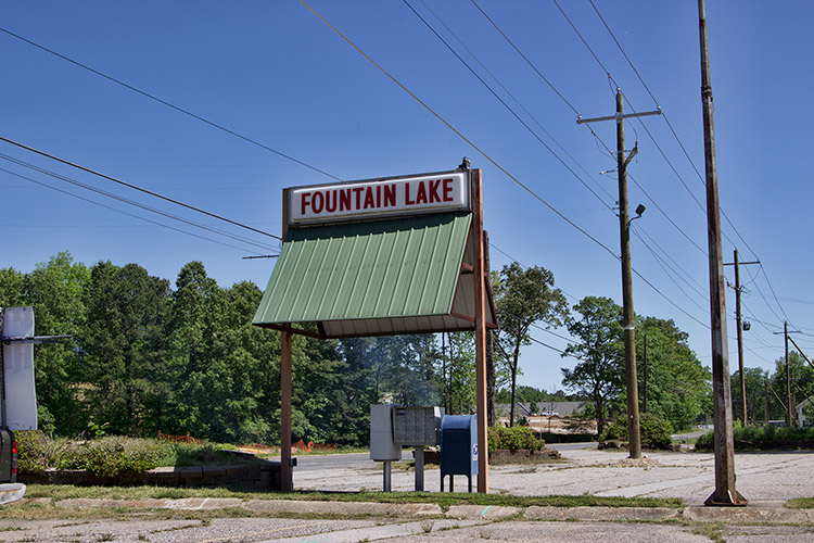 "Fountain Lake" sign with green awning over mailboxes in parking lot with power lines crossing above it