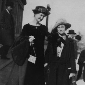 Two white women with hats and coats standing outside building