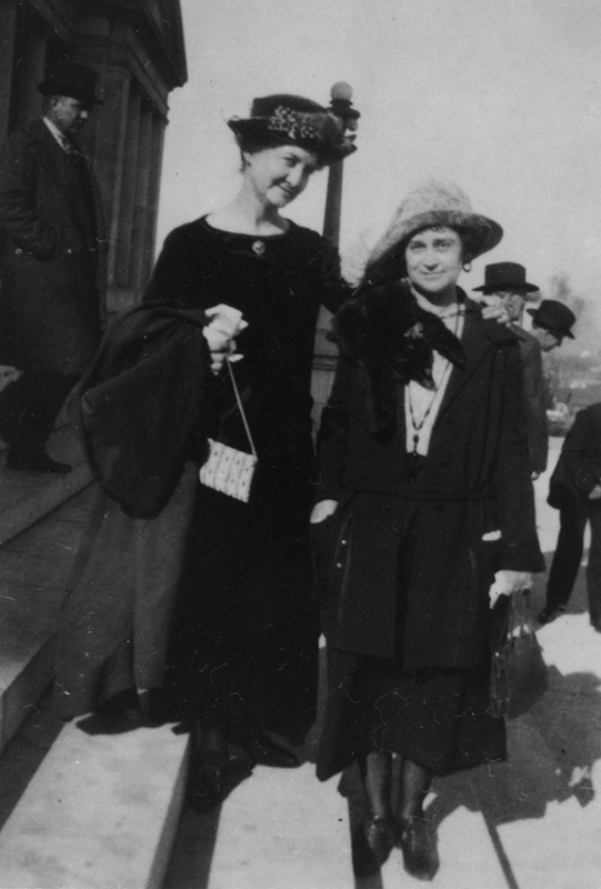 Two white women with hats and coats standing outside building