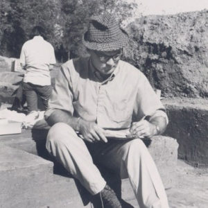 White man with hat and glasses taking notes on archaeological site with woman standing behind him