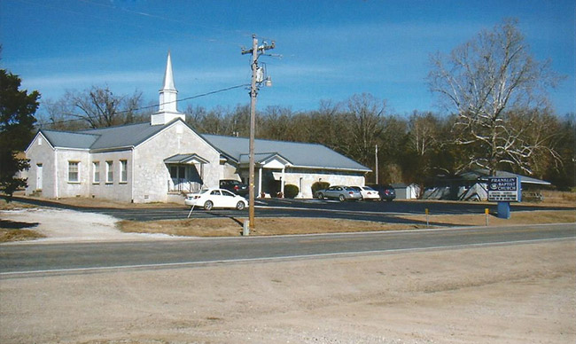 Single-story building with covered porch and steeple with parking lot and sign on street