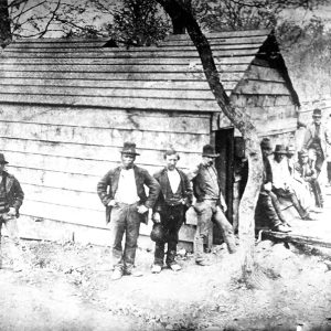 Group of white men with hats outside small building and tree