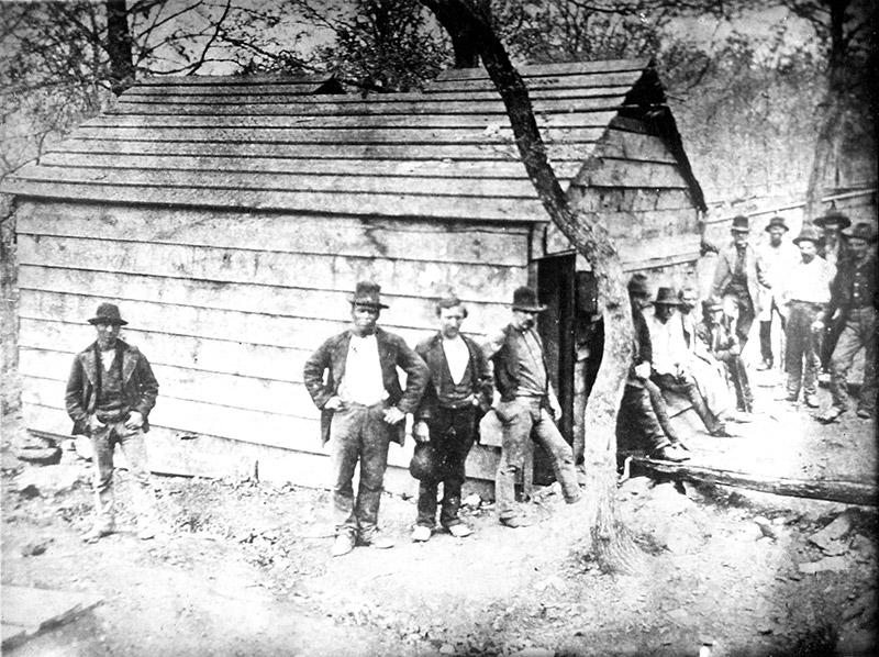 Group of white men with hats outside small building and tree