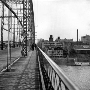 People walking across steel arch bridge over river with multistory buildings in the background