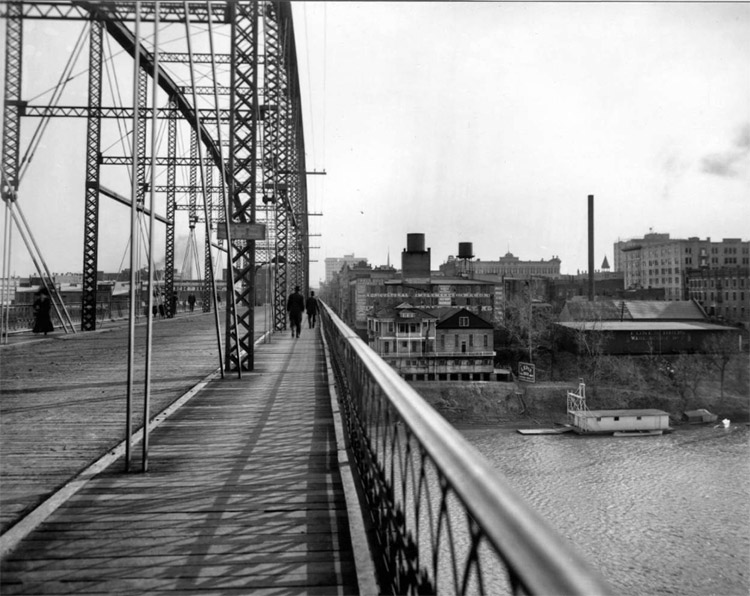 People walking across steel arch bridge over river with multistory buildings in the background