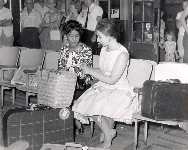African-American woman and white woman sitting indoors with their bags with white crowd behind them