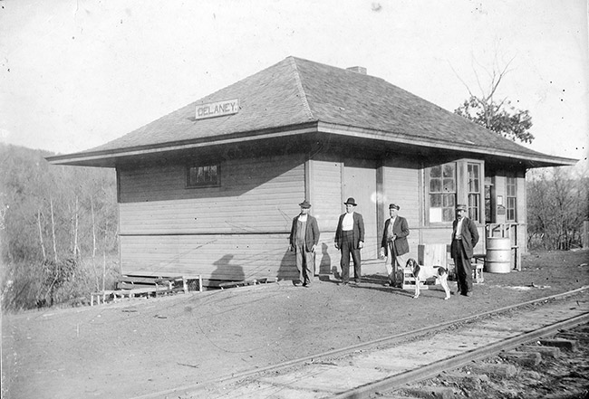 Four white men and dog at train depot building with tracks