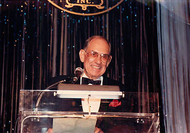 Old white man with glasses and suit smiling at lectern