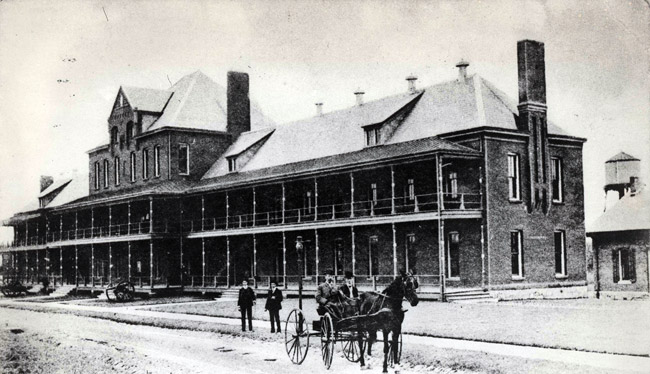 White men in suits and white men on horse drawn carriage outside long multistory building with covered porch and balcony