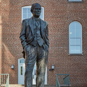 Statue of man in suit and bow tie on pedestal with multistory brick building behind it