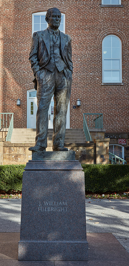 Statue of man in suit and bow tie on pedestal with multistory brick building behind it