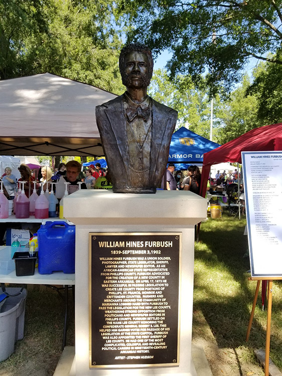 Bust of white man with beard in suit on pedestal with plaque on it and festival tents in background