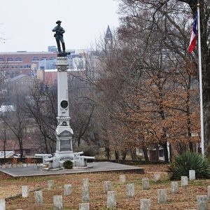 Tall stone monument with soldier on top and cannons around the bottom in cemetery with town buildings in the background